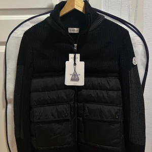 Moncler Cardigan - OBS: stretchy tung material, blivit stretchad där nere därav passar den bättre med en bredare kroppstyp. Svart stickad kofta från Moncler med quiltade partier framtill och två stora fickor. Hög krage, hel dragkedja och Moncler-logga på ärmen. Snygg mix av stickat och glansigt material, perfekt för kyliga dagar.