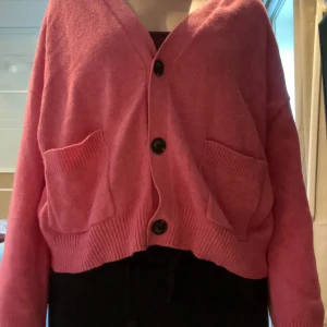 Rosa kofta med knappar och fickor - Mysig rosa kofta med v-ringning, stora svarta knappar och två rejäla fickor framtill. Koftan har ribbade muddar och en loose passform som ger en chill vibe. Perfekt att slänga på sig över en t-shirt eller linne.