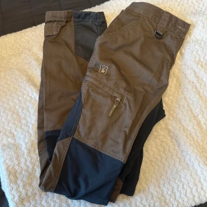 Deerhunter Lady Roja byxor brun/svart - Snygga Deerhunter Lady Roja byxor i brunt och svart med praktiska fickor och dragkedja. Perfekta för utomhusaktiviteter, tillverkade i slitstarkt material och med detaljer som D-ring och förstärkta partier.