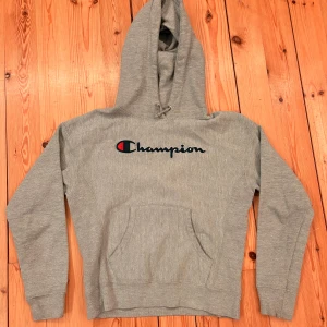 Grå hoodie från Champion XS - Grå hoodie från Champion i Reverse Weave-modell med broderad logga på bröstet och liten logga på ärmen. Klassisk känguruficka framtill, snörning i huvan och mjukt material. Perfekt för chill dagar eller streetwear-stil.