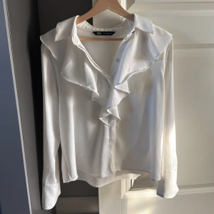 Vit volangblus från Zara - Säljer en vit skjorta från Zara med snygga volangdetaljer längs kragen och framtill. Skjortan har klassisk krage, knappar och långa ärmar. Materialet är mjukt och har en lätt glansig finish som ger en lyxig känsla.