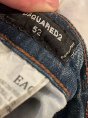 Dsquared2 blå slitna jeans herr - Säljer ett par blå jeans från Dsquared2 i storlek 52. Jeansen har en snygg tvätt med slitningar, färgstänk och distressed detaljer på benen. Klassisk femficksmodell och orange sömmar. Materialet är denim i bomull som ger en skön och trendig look. Knappast använd 