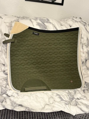 Olivgrön schabrak med vit kant - Säljer ett olivgrönt schabrak med vit kant och geometriskt mönster. Undersidan är svart med samma mönster. Schabraket har förstärkning vid sadelgjorden och praktiska fästen. Perfekt för dig som rider och vill ha stilren utrustning. Ej tvättad.