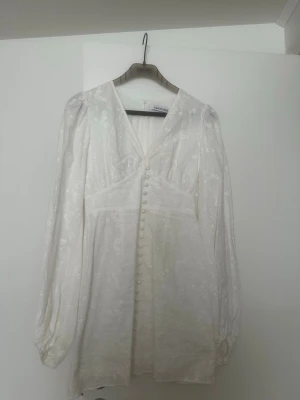 Vit blommig klänning från Adoore - Superfin vit klänning från Adoore med v-ringning, långa puffärmar och knappar framtill. Klänningen har ett subtilt blommigt mönster och är gjord i ett lätt tyg som känns lyxigt. Perfekt för sommarens alla tillfällen! Kan gå ner i pris vid snabb affär 