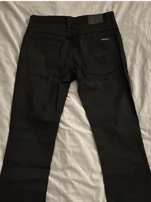 Svarta jeans från Nudie Jeans - Snygga svarta jeans från Nudie Jeans med klassisk femficksdesign och diskret logga på bakfickan. Jeansen har raka ben och är tillverkade i ett slitstarkt denimtyg. Perfekta för en stilren och enkel look.