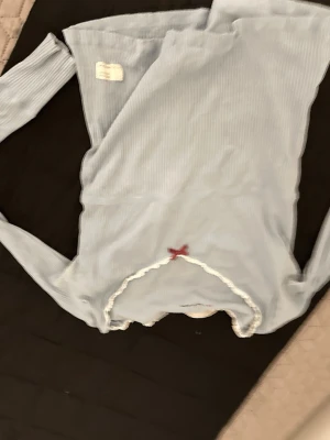 Ljusblå ribbad långärmad topp - Supersöt ljusblå långärmad topp med ribbad struktur och vit spetskant. Dekorerad med en liten röd rosett framtill. Perfekt för en trendig och söt look.