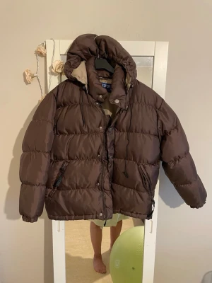  pufferjacka med huva - Snygg brun pufferjacka från SPORT med stor huva och dragkedja framtill. Jackan har quiltad design, långa ärmar och två sidofickor med dragkedja. Perfekt för kalla dagar och riktigt skön 