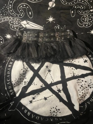 Svart kort tyllkjol med bältdetaljer - En svart kort kjol i tyll med bred midja och dubbla bältdetaljer med silverfärgade spännen och öljetter. Kjolen har en volangig och fluffig form som ger en edgy och alternativ vibe. Perfekt för dig som gillar statement-plagg och vill sticka ut.