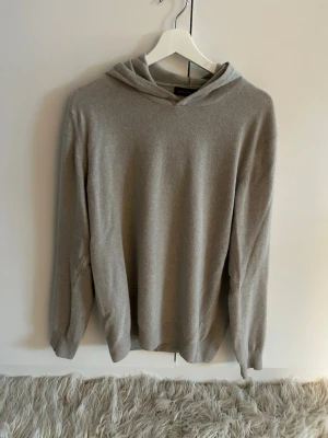 Beige Kashmir Hoodie Oscar Jacobsson - Säljer en stilren Beige hoodie från Oscar Jacobsson. Storlek M passar även S, hoodien är tillverkad i ett mjukt och tunt stickat material, modellen på bilden är cirka 183cm väger cirka 68kg, hoodien har långa ärmar och ribbade muddar vid ärmslut och nederkant. Perfekt för en avslappnad och snygg look.
