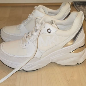 Vita sneakers från Michael Kors - Stilrena vita sneakers från Michael Kors med platåsula  och silvriga detaljer på hälen. Skorna har tydlig MK-logga på sidan, samt vita snören. Tillverkade i en mix av läder och syntetmaterial för en modern look.