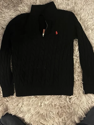 Svart kabelstickad tröja Polo Ralph Lauren - Kabelstickad svart tröja från Polo Ralph Lauren med halv dragkedja och hög krage. Tröjan har ett rött broderat logomärke på bröstet och ribbade muddar vid ärmslut och nederkant. Perfekt för lager på lager och snygg till jeans.