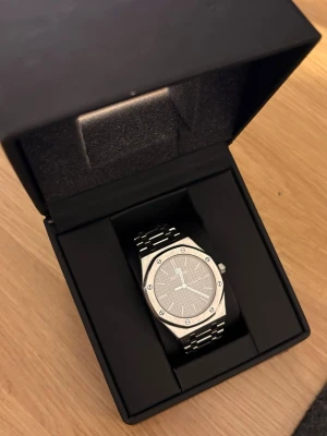 Silverfärgad klocka med länkarmband - Stilren klocka i silver med länkarmband och åttakantig boett. Urtavlan är grå med minimalistiska markeringar och visare. Klockan levereras i en svart box med glaslock, perfekt för förvaring eller present.