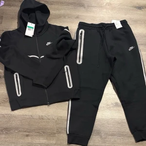 Svart Nike set med huva och byxa - Snyggt svart set från Nike med zip-hoodie och matchande byxor. Setet har vita och grå detaljer längs ärmar och ben samt Nike-logga på både tröja och byxa. Perfekt för en sportig och avslappnad look.