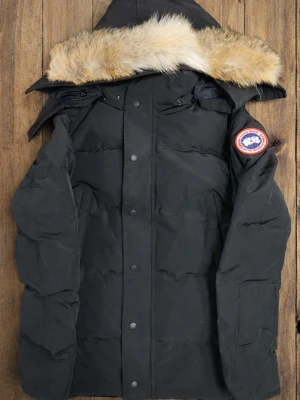 Svart Canada Goose Jacka - Snygg svart dunjacka från Canada Goose med avtagbar huva och äkta pälsdetalj. Jackan har klassisk logga på ärmen och stängs med både dragkedja och knappar. Perfekt för kalla vinterdagar och riktigt varm tack vare dunfyllningen.