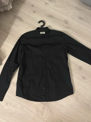 Svart skjorta från PRODUKT - Klassisk svart skjorta från produkt som tillhör Jack and Jones med lång ärm och knappar framtill. Skjortan har en stilren design och är tillverkad i ett mjukt bomullstyg som känns skönt mot huden. Perfekt för dig som gillar minimalistisk och tidlös stil.