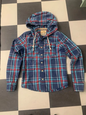 Rutig blå skjorta med huva Hollister - Rutig skjorta från Hollister i blått, rött och vitt med huva och snörning. Långärmad modell med knappar framtill och två bröstfickor. Perfekt för en avslappnad stil.