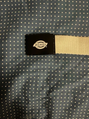 Vitt tygbälte från Dickies - Snyggt vitt tygbälte från Dickies med svart metallspänne och klassisk logga framtill. Perfekt för att ge din outfit en clean och stilren touch. Justerbar längd och slitstarkt material.