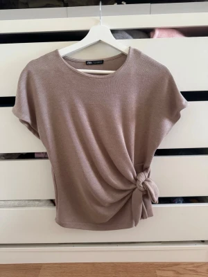 Beige topp med knyt från Zara - Stilren beige topp från Zara med korta ärmar och snygg knytdetalj i sidan. Perfekt för dig som gillar en enkel men trendig look. Mjuk och skön kvalitet.