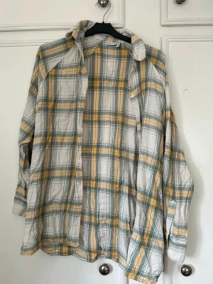 Rutigt overshirt i bomull - Luftig skjorta i bomull med rutigt mönster i vitt, gult och grönt. Skjortan har klassisk krage, knappstängning och en bröstficka. Perfekt för lager på lager och avslappnad stil.