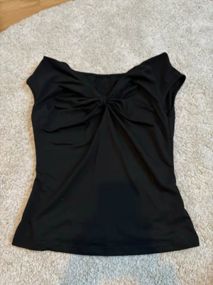 Svart topp med knutdetalj - Stilren svart topp med snygg knutdetalj framtill och bred halsringning. Modellen är ärmlös och har en figurnära passform som framhäver siluetten. Perfekt att matcha med jeans eller kjol för en trendig look.