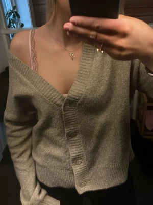 Beige kofta från Vero Moda - Mysig beige kofta från Vero Moda med djup V-ringning och stora knappar framtill. Koftan har ribbade muddar och en avslappnad passform som gör den perfekt att bära över ett linne eller t-shirt. Tillverkad i mjukt stickat material.