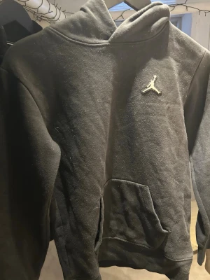 Svart hoodie från Jordan med logga - Säljer en svart hoodie från Jordan med den klassiska Jumpman-loggan broderad på bröstet. Hoodien har en stor känguruficka framtill och en skön huva. Tillverkad i mjukt bomullsmaterial som passar perfekt till chill eller streetwear.