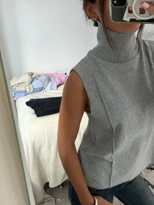 Grå ärmlös stickad polotröja - Säljer en stilren grå stickad polotröja utan ärmar. Tröjan har hög, ribbad polokrage och rak passform. Perfekt att styla med jeans eller kjol för en trendig look. Materialet känns mjukt och bekvämt mot huden.