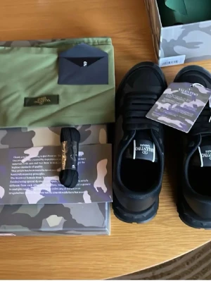 Svarta sneakers från Valentino Garavani - Snygga svarta sneakers från Valentino Garavani med diskret kamouflagemönster och logga på plösen. Skorna har klassisk låg profil, svarta skosnören och detaljer i läder och textil. Levereras med originalkartong, extra skosnören och dustbag.