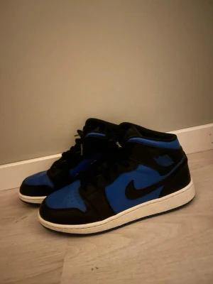 Air Jordan 1 Mid svart/blå - Snygga Air Jordan 1 Mid sneakers i svart och blått med klassisk Nike Swoosh på sidan. Skorna har en vit sula och är tillverkade i läder med perforerad tåbox och svarta skosnören. Perfekt för dig som gillar streetwear och vill sticka ut.