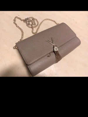 Beige/grå kuvertväska från Mario Valentino - Stilren beigegrå kuvertväska från Mario Valentino i läder med guldfärgad kedja och ikonisk V-logga framtill. Väskan har ett dekorativt tofs-hänge och ett rymligt fack med innerficka. Perfekt för att lyfta din outfit med en lyxig touch.