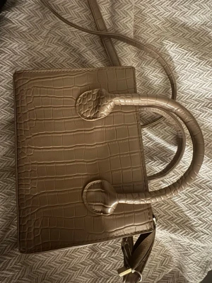 Beige handväska i krokopräglat material - Stilren beige handväska med krokodilpräglat mönster. Väskan har både handtag och axelrem för olika bärstilar. Rektangulär form och guldfärgade detaljer vid dragkedjan. Perfekt accessoar för en trendig look.