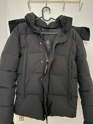 Svart dunjacka från Canada Goose - Snygg svart dunjacka från Canada Goose med avtagbar huva och diskret logga på ärmen. Jackan har dragkedja och knappar framtill samt två vertikala fickor. Perfekt för kalla vinterdagar och riktigt varm.