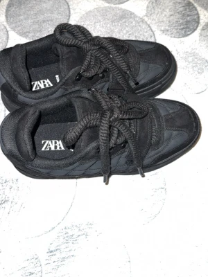 Svarta sneakers från Zara - Stilrena helsvarta sneakers från Zara med chunky sula och breda skosnören. Ovandelen är i en mix av textil och mockaimitation med diskreta sömdetaljer. Låg modell som passar perfekt till streetwear och casual outfits.