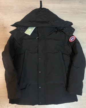  Canada Goose - Svart Canada Goose jacka röd logo i storlek S.      Jackan har huva, knappar framtill och en clean, stilren look. Perfekt för kalla dagar med sin tjocka och värmande fyllning av dun och fjädrar. Den är använd ett fåtal gånger men annars het ny så lappen är inte kvar längre. Påsen och pälsen följer med.