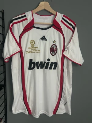 AC Milan Kaka' 22 Champions League tröja - Vit AC Milan matchtröja från Adidas med röda och svarta detaljer, Champions League-brodyr, Bwin-logga och klubbmärke fram. På ryggen står KAKA' och nummer 22. Tillverkad i lätt och ventilerande polyester, perfekt för fotbollsträning eller samling. Helt ny med tagg en replika 