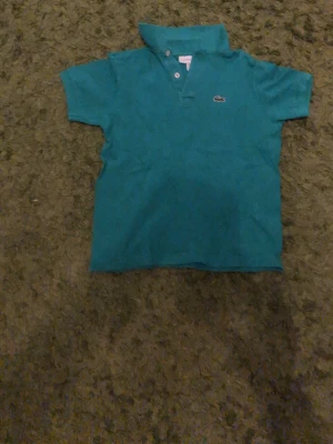 Turkos grön pikétröja från Lacoste - Klassisk turkos pikétröja från Lacoste med korta ärmar och den ikoniska krokodilloggan på bröstet. Tröjan har krage och knäppning framtill, perfekt för en avslappnad men ändå stilren look.