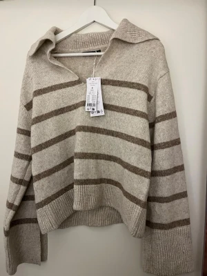 Beige stickad tröja med bruna ränder - Säljer en beige stickad tröja med bruna ränder. Tröjan har en bred krage, v-ringning och långa ärmar med ribbade muddar. Lappen är kvar