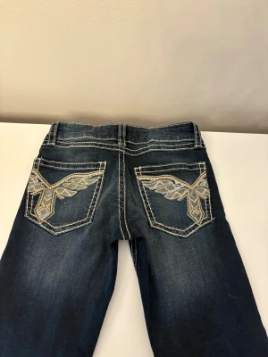Mörkblå jeans  - Snygga mörkblå jeans med kontrastsömmar och broderade detaljer i silver och guld på bakfickorna. Klassisk femficksmodell med bälteshällor och låg midja. 