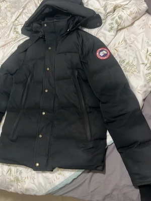 Svart dunjacka från Canada Goose - Säljer en svart dunjacka från Canada Goose med klassisk logga på ärmen. Jackan har huva, dragkedja och tryckknappar framtill samt två stora fickor med dragkedja. Perfekt för kalla vinterdagar och riktigt varm.