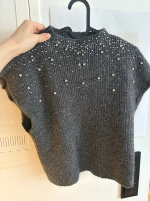 Grå stickad väst med pärlor - Säljer en grå stickad väst med glittriga pärlor upptill. Västen har en rak passform, ribbstickad struktur och är ärmlös. Perfekt att styla över en skjorta eller långärmad tröja för en trendig lager-på-lager-look.