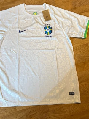 Vit Brasil Nike t-shirt med mönster - Säljer en vit Brasil t-shirt från Nike med diskret leopardmönster i tyget och blå Nike-logga på bröstet. T-shirten har korta ärmar med gröna och gula detaljer vid ärmsluten samt Brasil-märke på bröstet. Perfekt för dig som älskar fotboll och vill sticka ut.