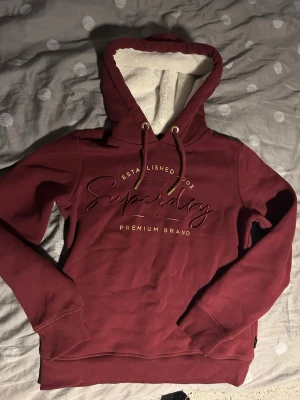 Vinröd hoodie från Superdry - Snygg vinröd hoodie från Superdry med broderad logga och text på bröstet. Hoodien har en fodrad huva med vita detaljer och dragsko med guldfärgade metalldetaljer. Mjuk och skön bomullsblandning, perfekt för en avslappnad stil.