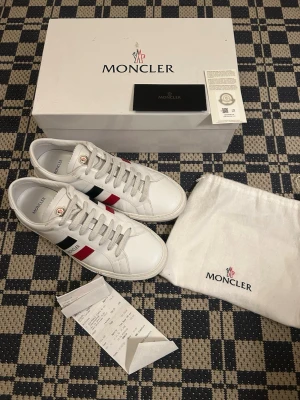 Vita Moncler sneakers med röd och blå detalj - Snygga vita sneakers från Moncler i läder med klassiska ränder i rött och blått på sidan samt Moncler-logga på tungan. Skorna har vita snören och en låg siluett som passar till många stilar. Kommer med originalkartong och dustbag.
