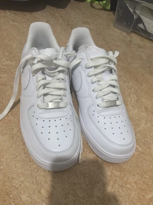 Vita Nike Air Force 1 sneakers - Klassiska helvita Nike Air Force 1 sneakers i lågt utförande. Skorna har en stilren design med perforerad tåbox, vita skosnören och den ikoniska swoosh-loggan på sidan. Tillverkade i slätt läder med en rejäl gummisula för skön komfort.