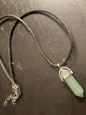 Halsband med grön kristallhänge - Snyggt halsband med ett grönt kristallformat hänge i sten och silverfärgad infattning. Hängs i ett svart, flätat band med justerbar kedja. Perfekt accessoar för dig som gillar kristaller och stilrena smycken.