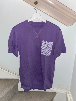 Lila Adidas t-shirt med mönstrad ficka - Lila t-shirt från Adidas med korta ärmar och en vit-lila mönstrad bröstficka. Klassisk rund halsringning och diskret Adidas-logga på bröstet. Perfekt för en avslappnad och sportig stil. Storlek S