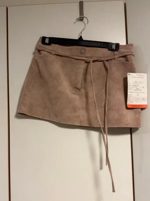 Beige mockakjol med knyt från H&M - Säljer en beige kortkjol i mockaimitation från H&M. Kjolen har knytband i midjan, bälteshällor och knapp framtill. Storlek 38 men passar även 36. Denna är från en provkollektion som har hängt ute på en mässa