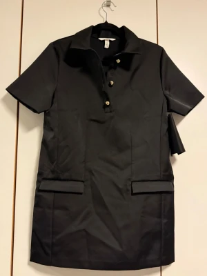 Svart skjortklänning med fickor - Stilren svart skjortklänning med korta ärmar och krage. Klänningen har tre knappar framtill och två riktiga fickor vid höfterna. Rak passform. Denna är troligtvis från en provkollektion som har hängt ute på en mässa