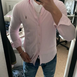 Ralpha lauren skjorta  - Snygg rosa randig skjorta från Ralph Lauren med klassisk krage och broderad logga på bröstet. Skjortan har långa ärmar och knappar framtill. Perfekt för en stilren look.