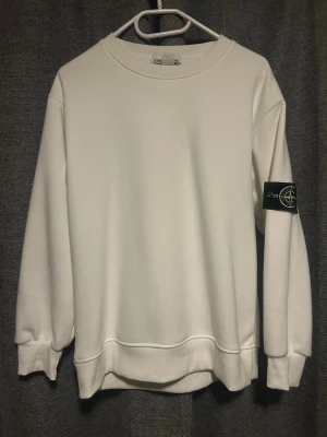 Vit sweatshirt från Stone Island - Säljer en clean vit sweatshirt från Stone Island med den klassiska loggan på vänster ärm. Tröjan har rund halsringning, ribbade muddar och en relaxed passform. Perfekt för dig som gillar stilrena plagg med streetwear-vibe.
